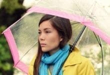 mujer de aspecto triste con un abrigo y una bufanda parada debajo de un paraguas en la lluvia