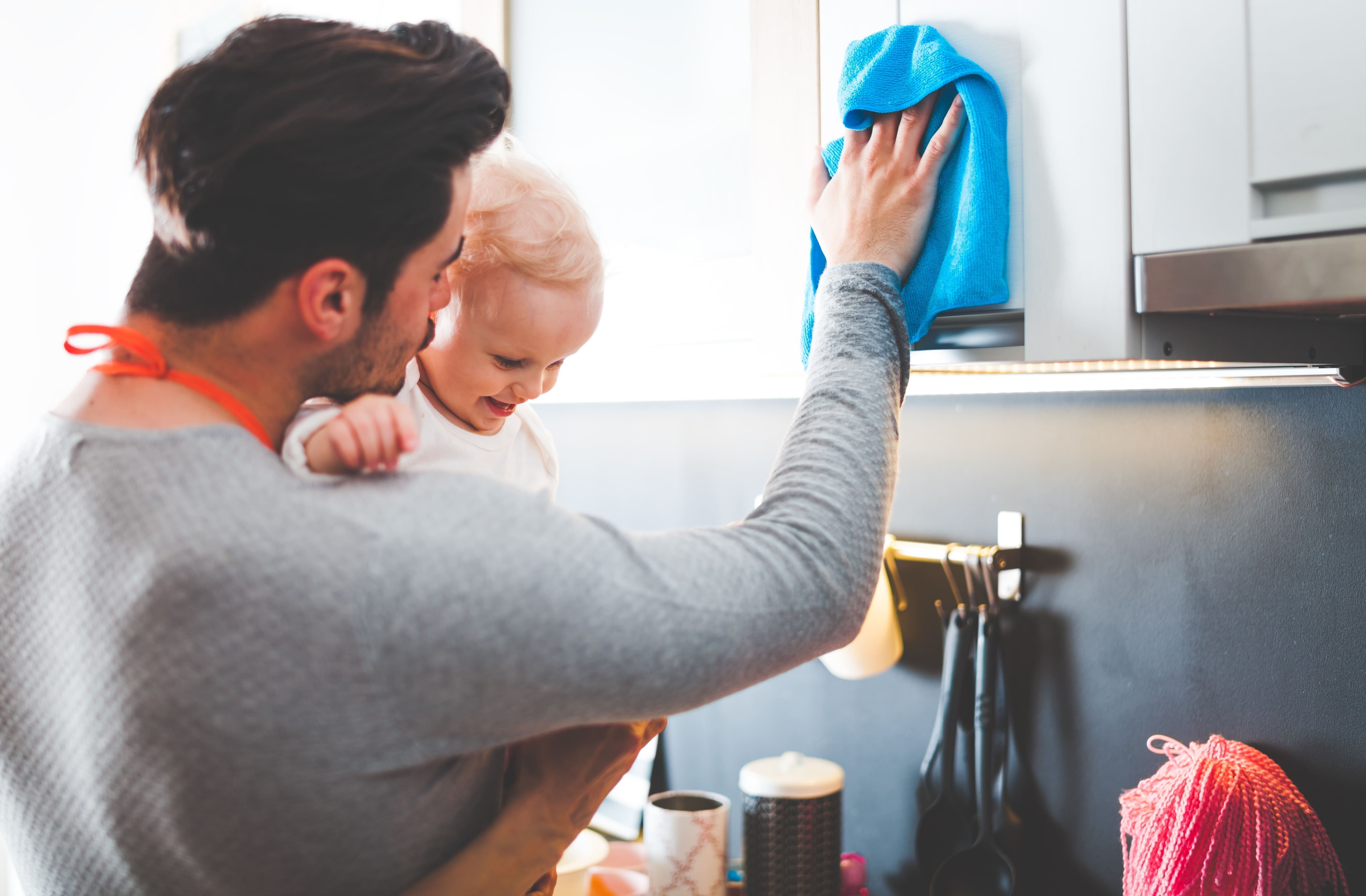A young father cleans the kitchen while holding his daughter.